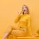 Woman sitting while wearing a yellow outfit against a yellow background.
