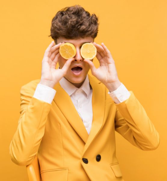 Homme en costume jaune tenant deux moitiés de citron devant ses yeux sur fond jaune.