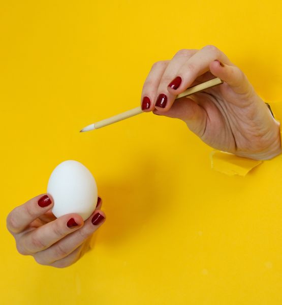Hand with red nail polish holding a white egg and a paintbrush against a bright yellow background.