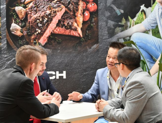 Four professionals engaged in a discussion around a table at a trade show, with a large grilled meat visual in the background.