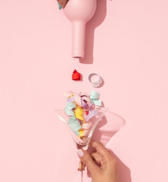 Hand holding a martini glass filled with colorful ribbons, with a pink bottle positioned upside down above it on a pink background