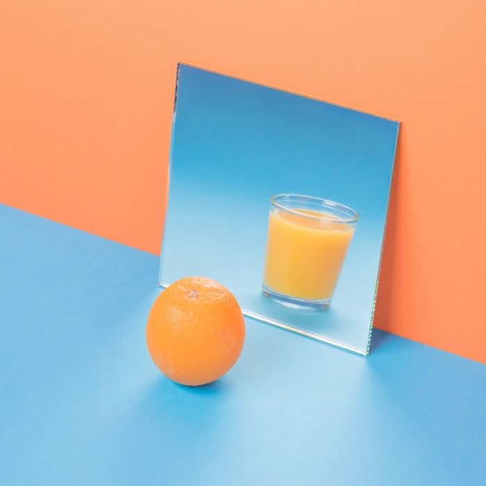Orange entière posée devant un miroir reflétant un verre de jus d’orange sur fond coloré