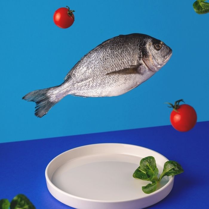 Poisson entier suspendu au-dessus d’une assiette blanche avec tomates et herbes