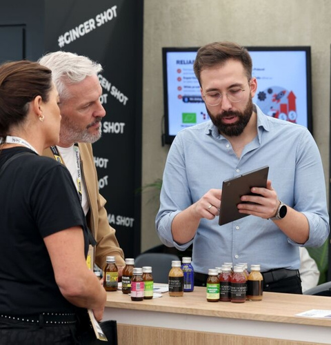 Man explaining beverage products to two visitors at a trade show, holding a tablet at the booth.