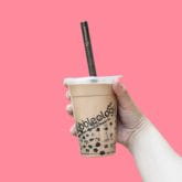 A hand holds a bubble tea with black pearls, against a pink background.