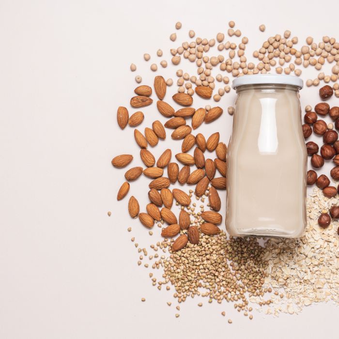 Bouteille de lait végétal entourée de graines de soja, amandes, noisettes et quinoa sur fond beige.