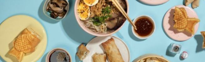 Top view of an assortment of Asian dishes including noodles, spring rolls, gyozas, steamed buns, and fortune cookies.
