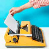 Main insérant une feuille de papier dans une machine à écrire jaune, avec un fond bleu.