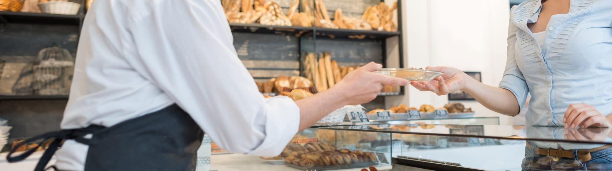 Bakery staff member serving a customer beside a pastry display case and shelves filled with artisan breads.