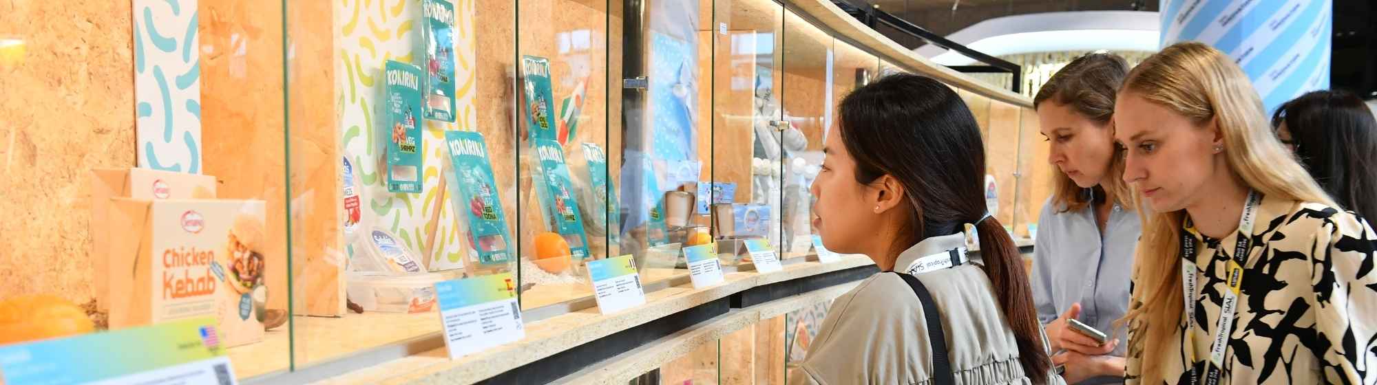 Three women closely observe food products displayed behind a glass showcase.