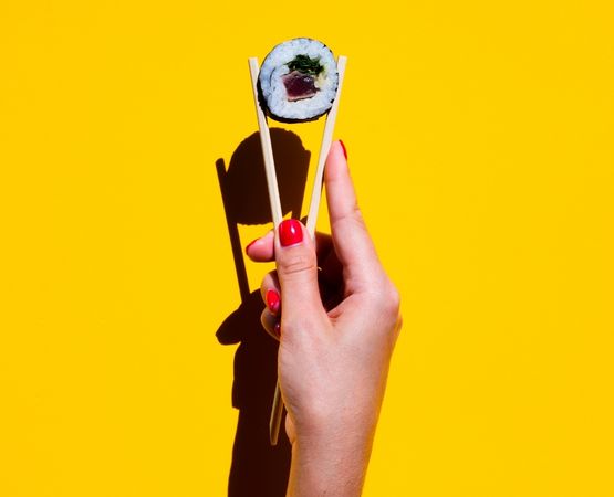 Hand holding a maki roll with chopsticks on a bright yellow background, illustrating Japanese food.