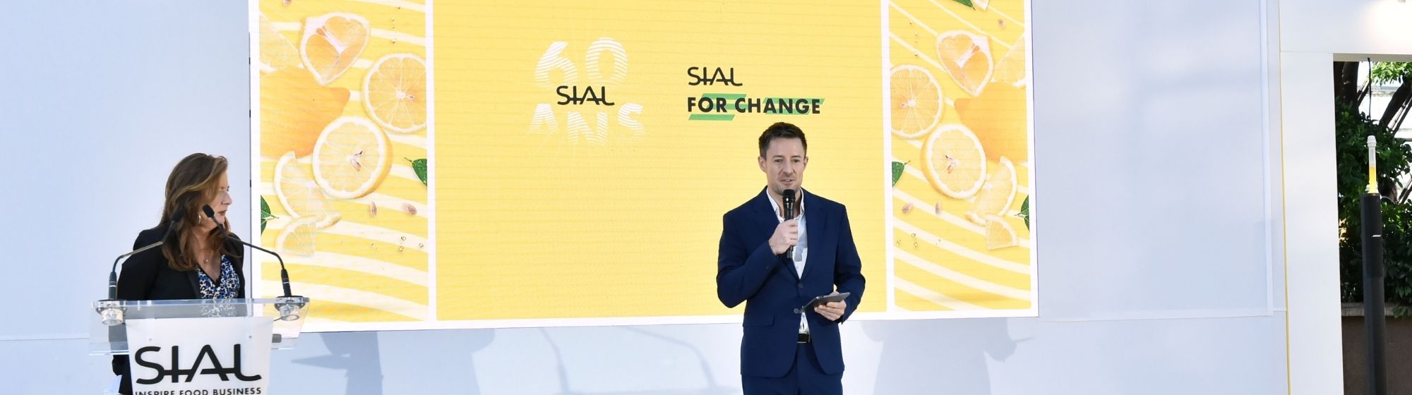 Speaker with microphone on the SIAL For Change stage, with yellow screen and SIAL podium.