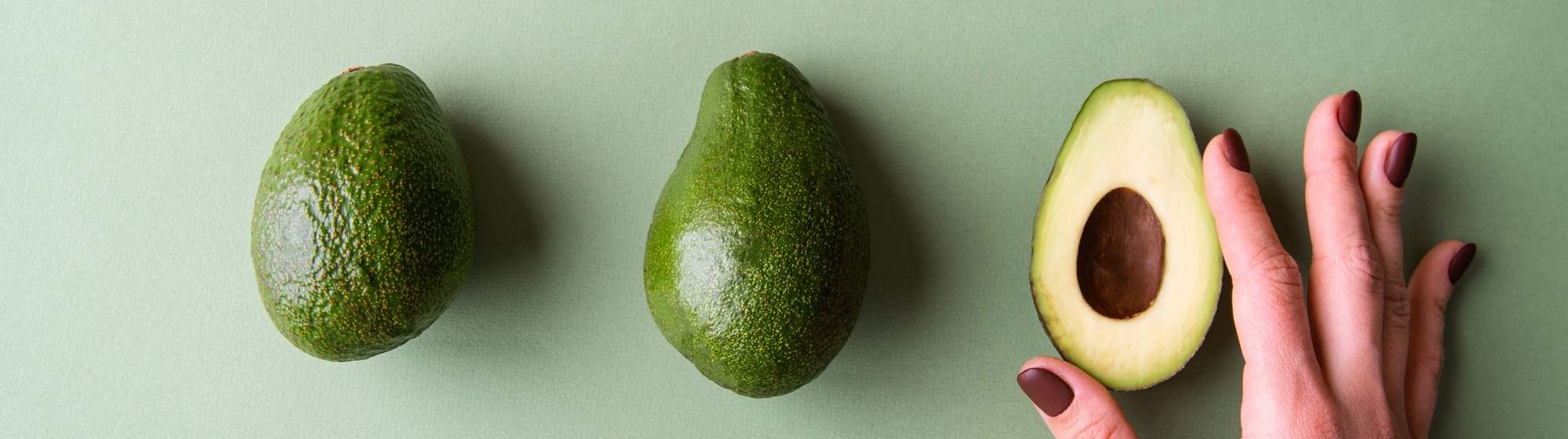 Banner with two whole avocados, one avocado half and a hand on a light green background.