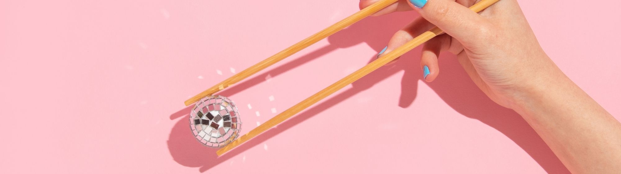 Main avec baguettes tenant une petite boule disco sur fond rose minimaliste, visuel artistique et moderne inspiré pop culture