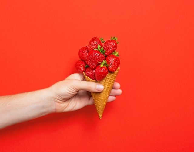 Hand holding a cone filled with fresh strawberries on a red background.