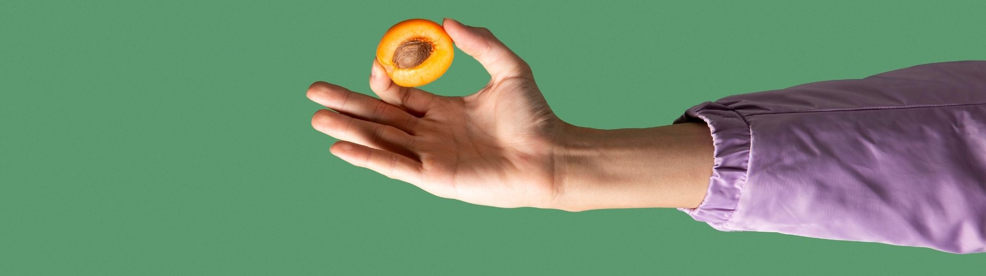 Extended arm holding a halved apricot with pit on a green background.