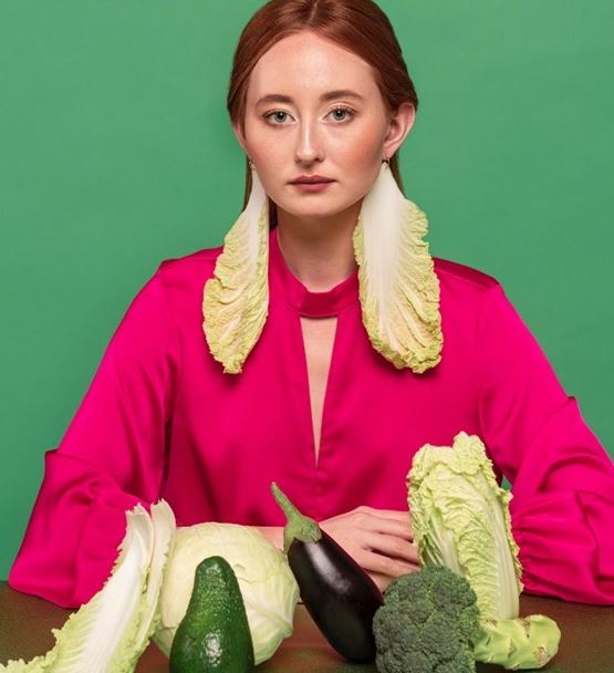 Woman wearing cabbage leaves as earrings, surrounded by vegetables, green background.