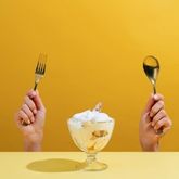 Ice cream cup held between fork and spoon on yellow background, concept of sweet food indulgence