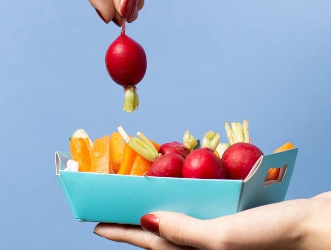 Hand holding a box of raw vegetables with radishes and veggie sticks on blue background, healthy snack concept