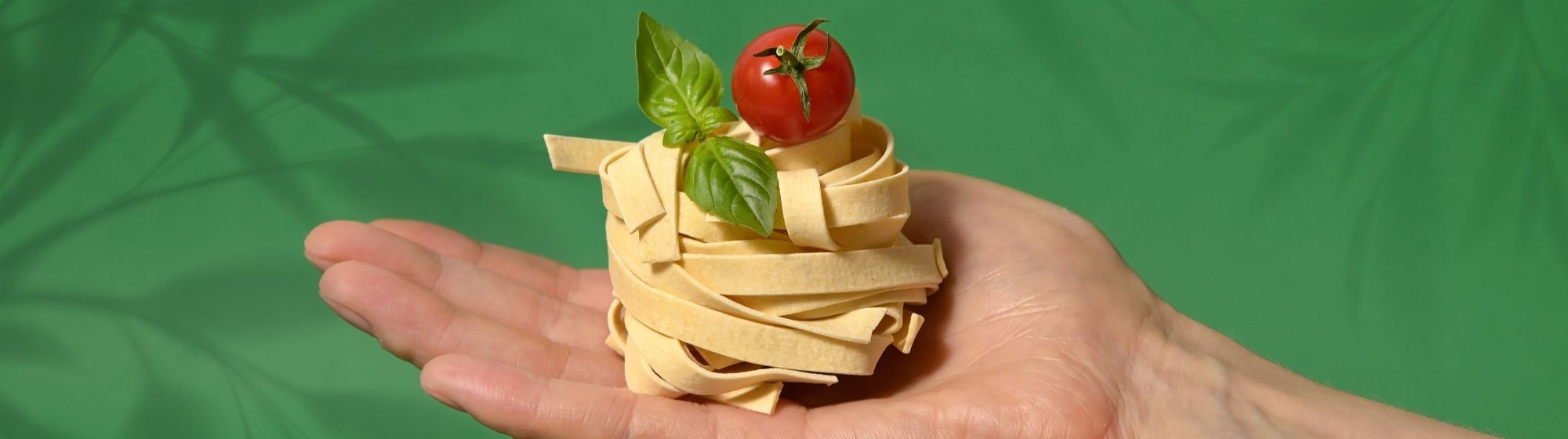 Stacked raw tagliatelle pasta with cherry tomato and basil leaves on a green background