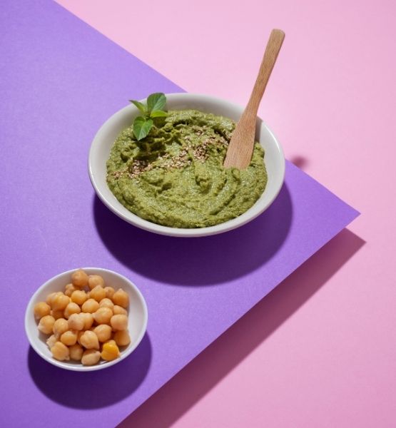 Bowl of green hummus with a wooden spoon and chickpeas on a pink and purple background.