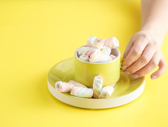 Yellow cup filled with marshmallow candies on a saucer, with a hand on a yellow background.