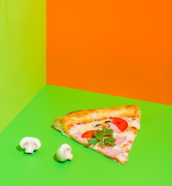 Slice of ham and mushroom pizza on green and orange background with fresh mushrooms.