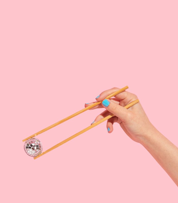 Hand holding a small disco ball with chopsticks against a pastel pink background.