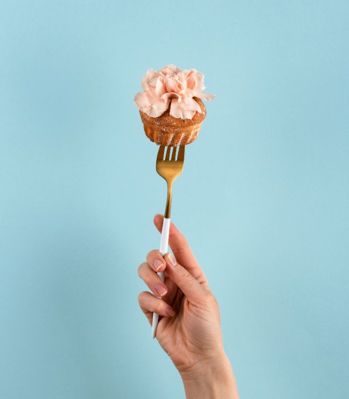 Main tenant une fourchette dorée piquée dans un cupcake décoré de pétales roses, sur fond bleu clair.