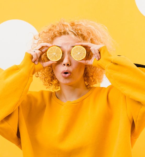 Woman wearing a yellow sweater holding two lemon halves over her eyes against a yellow background with white dots.