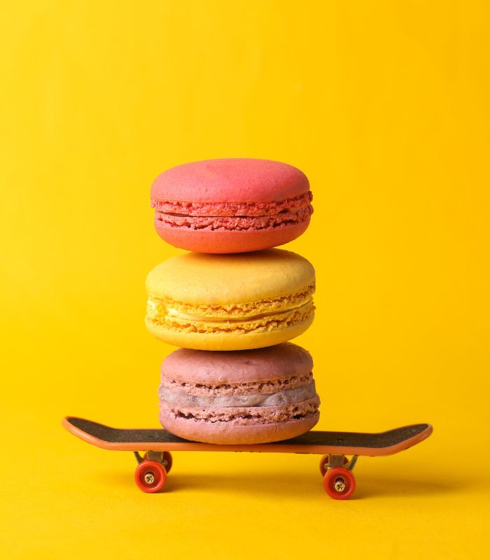 Trois macarons colorés empilés sur un mini-skateboard, sur fond jaune vif.