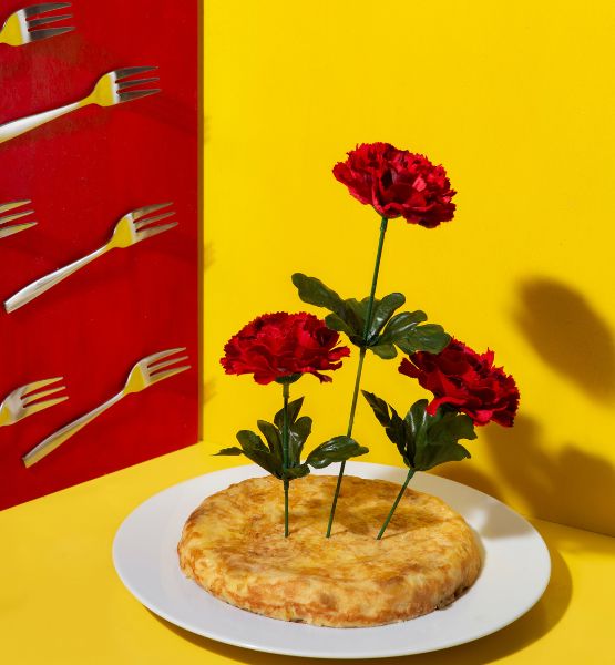 Omelette posée sur une assiette blanche avec des fleurs rouges plantées au centre, sur fond jaune, avec des fourchettes fixées sur un panneau rouge.