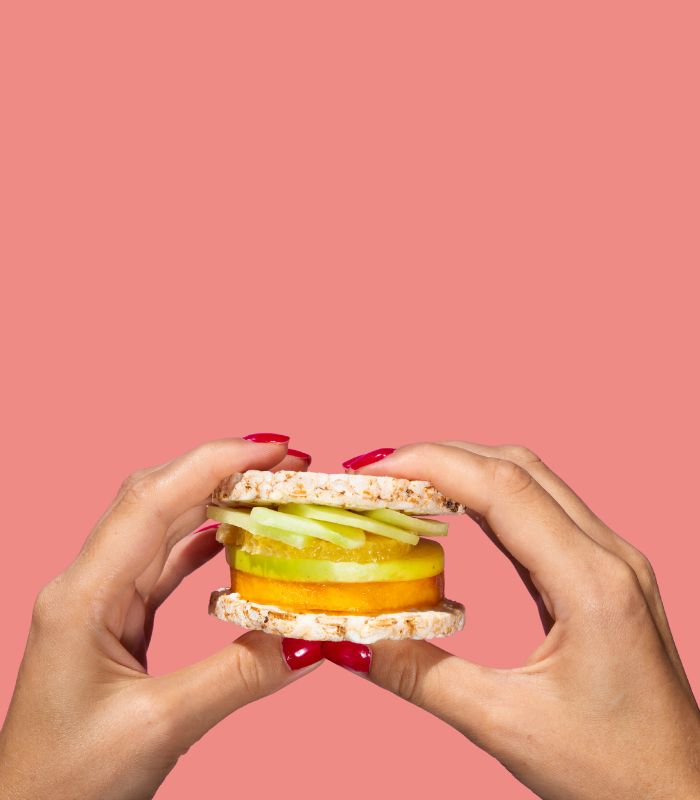 Hands holding a sandwich made of rice cakes and fruit slices, against a salmon-pink background.