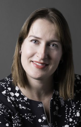 Portrait professionnel d’une personne souriante portant un haut noir à motifs blancs sur fond gris sombre.