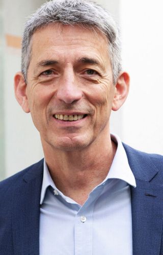 Portrait professionnel d’une personne souriante aux cheveux gris, portant une veste bleue et une chemise claire.